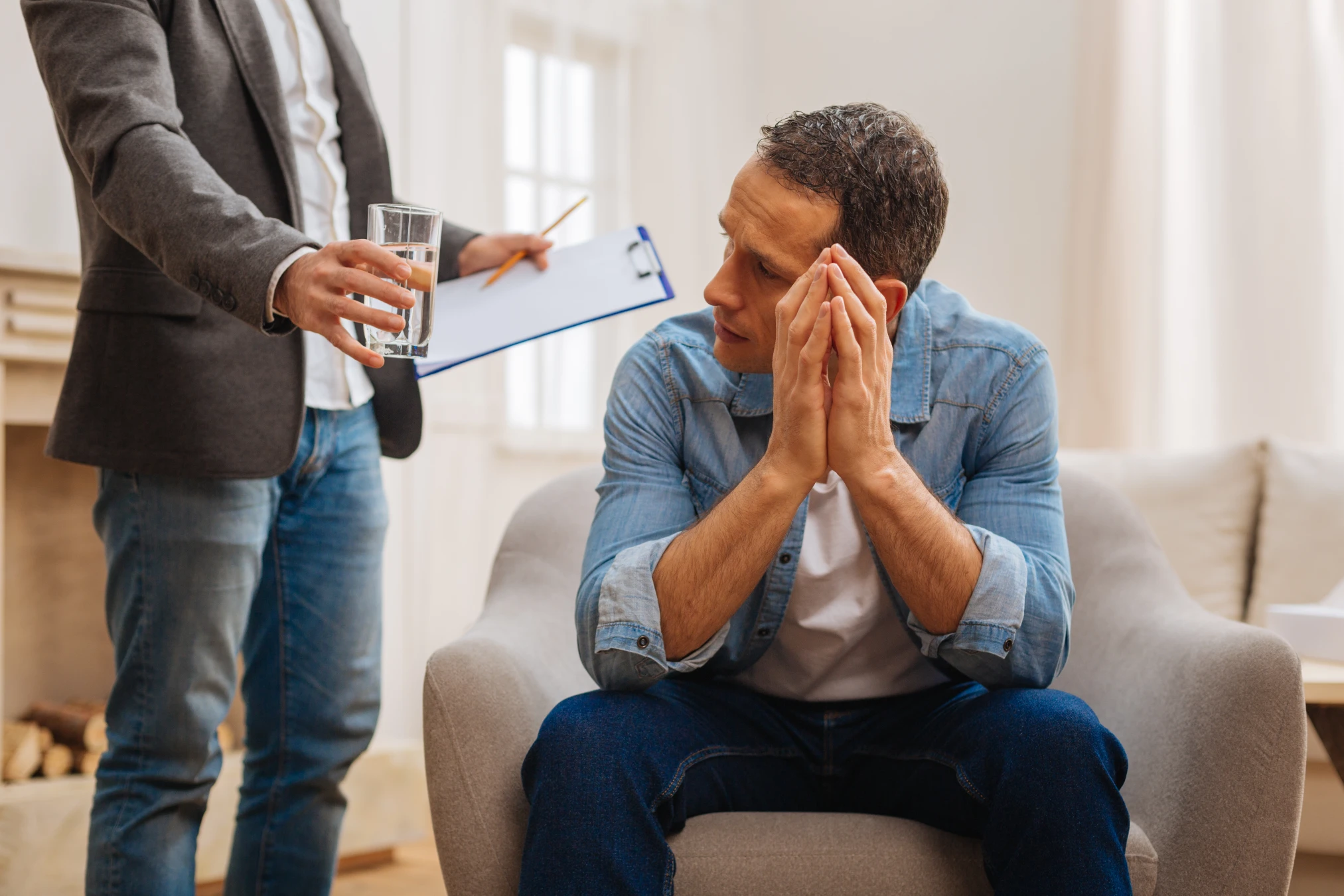 a concerned man sitting in a chair recieves a glass of water from another man
